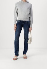 Personne portant un sweat à capuche gris clair, un jean bleu foncé, des sandales blanches à talons, tenant un petit sac à main blanc sur un fond uni.