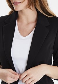 Svart kavaj med en strukturerad design, med slagkrage och trekvartslånga ärmar, över en figurnära vit t-shirt med v-ringning.