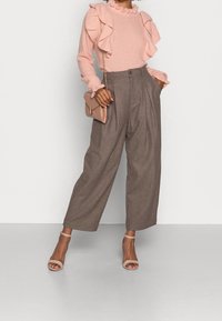 Světlé růžové svetr s texturou a volánovými akcenty v kombinaci s hnědými kalhotami s vysokým pasem a širokými nohavicemi. Součástí je malá béžová kabelka.