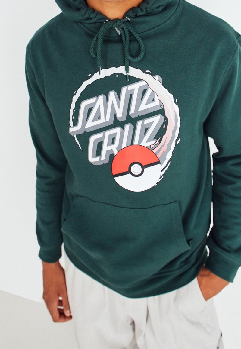 Persona que lleva una sudadera con capucha de color verde oscuro con el texto "Santa Cruz", un gráfico de una ola en círculo y un diseño de Poké Ball en rojo y blanco en la parte delantera.