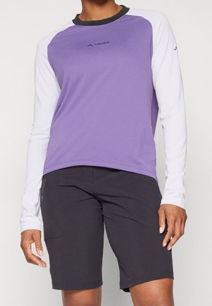Person trägt ein lila-weißes langärmeliges Sportoberteil und schwarze knielange Shorts vor einfachem Hintergrund.