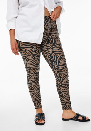 Bruine en zwarte zebraprint legging met een fitted design, gecombineerd met zwarte slide sandalen met metalen accenten. Witte button-up shirt.