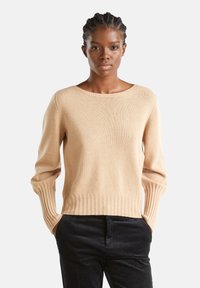 Frau mit geflochtenem Haar, die einen beigen Strickpullover und schwarze Hosen trägt, mit den Händen in den Taschen stehend, vor einem weißen Hintergrund.