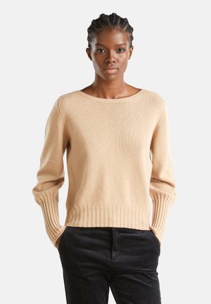 Femme aux cheveux tressés portant un pull en tricot beige et un pantalon noir, se tenant les mains dans les poches contre un fond blanc.