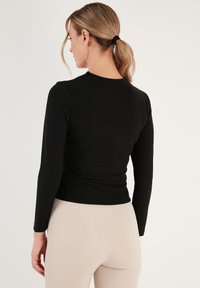 Top nero a maniche lunghe con texture a coste e design aderente, con scollatura tonda e orlo posteriore dritto. Capelli raccolti visibili.