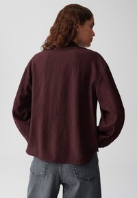 Bordeauxkleurig shirt met textuur, lange mouwen, een ontspannen pasvorm, een afgeronde zoom en een subtiel gekreukt patroon, gecombineerd met grijze spijkerbroek.