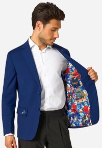 Vīrietis ziliem žaketē un baltos kreklos atver žaketi, lai parādītu iekšējo daļu ar krāsainiem Sonic the Hedgehog tēla zīmējumiem.