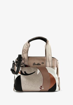 Sac à main beige et marron avec des poignées beiges à motif, une bandoulière noire ajustable et un design abstrait de visage avec de petits détails cloutés sur la poche avant.
