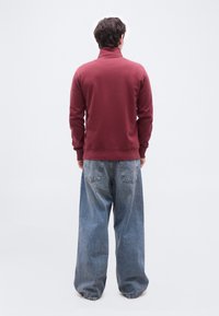 Άνδρας με την πλάτη γυρισμένη, φοράει μπορντό φούτερ με λαιμόκοψη και φαρδιά ξεθωριασμένα μπλε τζιν, μπροστά σε απλό λευκό φόντο.