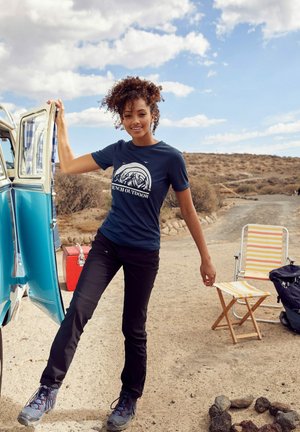 Navy T-Shirt mit einem Berggrafik und dem Text "French Outdoor", kombiniert mit schwarzen Hosen und grauen Wanderschuhen, vor einem blauen Van.