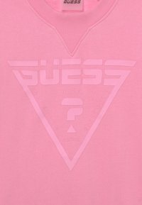 Maglione rosa con un logo GUESS triangolare testurizzato, con "NINETEEN", "EIGHTY-ONE" e "ACTIVE" stampati lungo i bordi.
