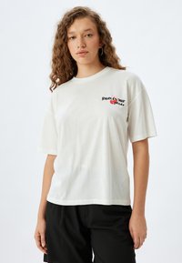 Camiseta de algodón blanca de manga corta. Presenta un diseño gráfico de texto en negro y rojo en el lado izquierdo del pecho. Textura suave, corte holgado.