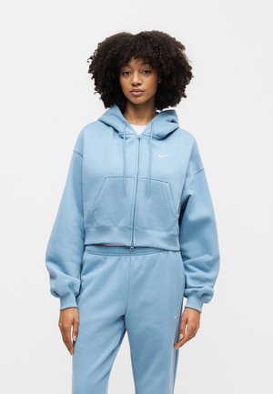 Kvinde med krøllet hår iført lyseblå Nike lynlås-hættetrøje og matchende joggingbukser, stående mod en ensfarvet lys baggrund.
