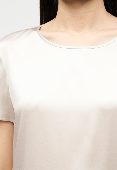 Γυναίκα που φοράει μπεζ σατέν τοπ με κοντά μανίκια και στρογγυλή λαιμόκοψη, εμφανιζόμενη από τον λαιμό έως τη μέση σε λευκό φόντο.