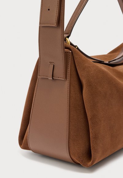Καφέ τσάντα χειρός από σουέντ και δέρμα με φαρδιά δερμάτινη λουρίδα και χρυσούς μεταλλικούς λεπτομερείς, εμφανιζόμενη από το πλάι με ορατά σημεία ραφής.