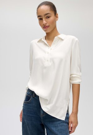 Junge Frau mit zurückgebundenem Haar, die eine weiße Langarmbluse und blaue Jeans trägt, steht vor einem schlichten hellen Hintergrund.