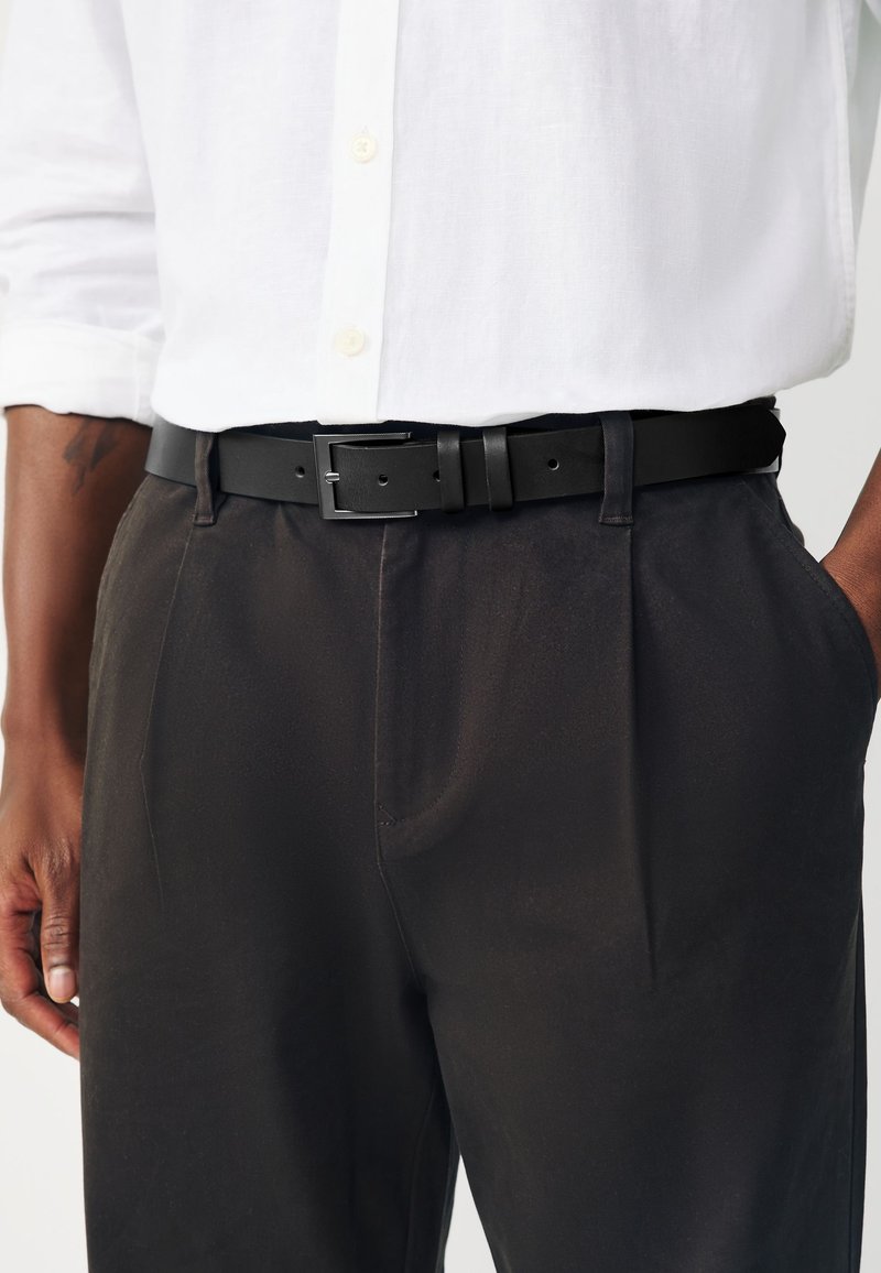 Personne portant une chemise blanche boutonnée rentrée dans un pantalon foncé avec une ceinture en cuir noire, une main dans la poche, tatouage visible sur l'avant-bras.