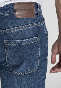 Traperice u tamnoplavoj boji s smešnim kožnim oznakom "GIANNI KAVANAGH" na zadnjoj strani. Imaju kontrastne šavove i stražnji džep.
