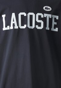 Tamno plava pamučna majica sa podebljanim bijelim slovima koja ispisuju "LACOSTE" te malim izvezenim krokodilom na prednjoj strani.