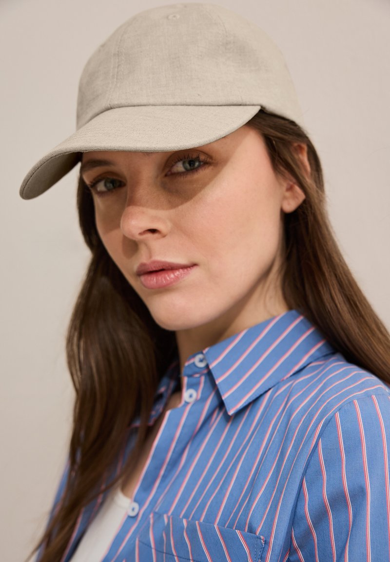 Gorra de béisbol beige con una textura suave, llevada ligeramente inclinada. La persona viste una camisa de rayas azul con líneas rosas.