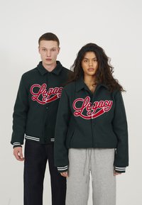 Mørkegrøn varsity jakke med stort rødt "Legacy" broderi, med kravehals, knapper og stribede manchetter.