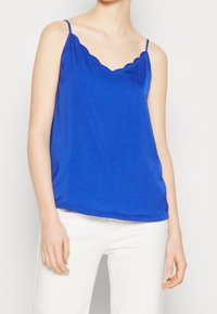 Camisole bleu roi avec encolure festonnée et fines bretelles, fabriquée dans un tissu lisse. La silhouette est ample et légèrement drapée.