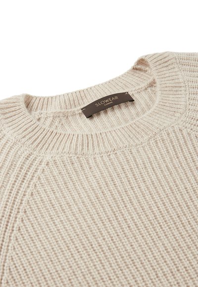 Cremfarbener Strickpullover mit gerippter Textur, der einen runden Ausschnitt aufweist und ein Etikett mit der Aufschrift "SLOWEAR" in einer kontrastierenden dunklen Farbe trägt.