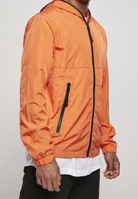 Orange letvægtsjakke med hætte, sort lynlås, elastiske manchetter og sidelommer; lavet af glat, tyndt stof med en let glans.