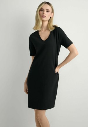 Kobieta o blond włosach do ramion, ubrana w czarną sukienkę z dekoltem w kształcie litery V, krótkimi rękawami i długością do kolan, z kieszeniami, stojąca na jednolitym jasnym tle.