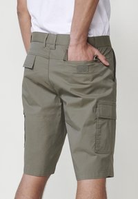 Uomo che indossa pantaloni cargo verde oliva con più tasche, mano parzialmente nella tasca posteriore, abbinati a una maglietta bianca.