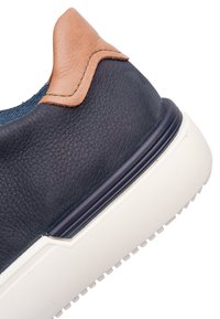 Detailní pohled na patu tenisky z tmavě modré kůže s béžovým koženým poutkem a silnou bílou gumovou podrážkou.