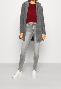 Abrigo gris de espiga con capucha, combinado con un suéter burdeos y unos jeans skinny gris claro. Zapatillas blancas completan el atuendo.