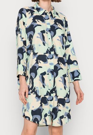 Nainen, joka käyttää löysää, pitkähihainen paitamekkoa, jossa on kauluksellinen pääntie ja abstrakti sininen, musta ja beige kuvio.