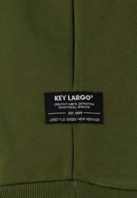 Tessuto verde oliva con una superficie liscia, caratterizzato da un'etichetta nera che riporta la scritta "KEY LARGO" e ulteriore testo bianco.