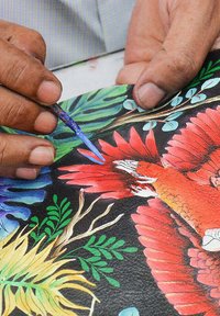 Een hand houdt een fijne kwast die levendige bloemen- en vogelontwerpen in rood, groen en blauw op textuur zwart materiaal schildert.