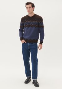 Stickad tröja i blått och brunt med geometriskt mönster, rund halsringning, ribbad mudd, kombinerad med blå jeans och svarta skor.