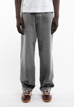 Personne portant un jean gris ample et des baskets marron avec des lacets blancs, debout devant un fond blanc.