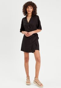 Schwarzes Button-Down-Kleid mit kurzen Ärmeln, Wickeldesign und seitlichem Bindegurt, aus leichtem Stoff; getragen mit beigen Riemchensandalen.