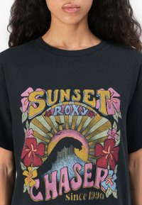 T-shirt noir avec un design graphique coloré incluant des accents floraux, des rayons de soleil et une vague. Le texte indique "SUNSET ROXY CHASER depuis 1990."