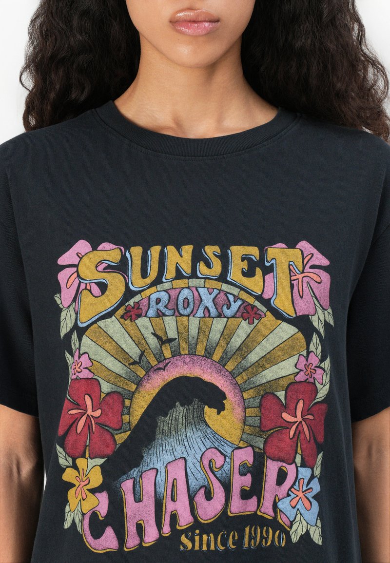 Czarna koszulka z kolorowym wzorem graficznym, z kwiatowymi akcentami, promieniami słońca i grafiką fal. Napis brzmi "SUNSET ROXY CHASER od 1990".