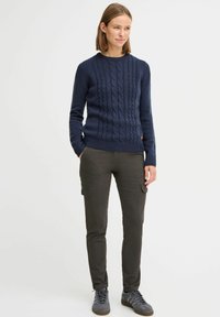 Marine blå kabelstrikket genser med rund hals, kombinert med mørke cargobukser med sidelommer. Modellen har på seg grå joggesko.