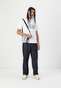 T-shirt gráfica branca com ilustração azul e verde, jeans escuros, mala de tiracolo azul clara e ténis bege.