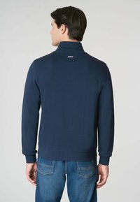 Giacca pullover navy con colletto alto, polsini e orlo a coste; presenta una piccola toppa con logo sul retro. Tessuto a maglia testurizzato.