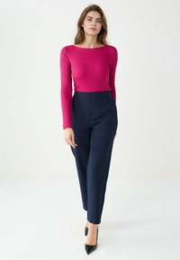 Getop met lange mouwen in felroze, gecombineerd met hooggetailleerde marineblauwe broek. Model draagt zwarte spits toelopende hakken, tegen een effen achtergrond.