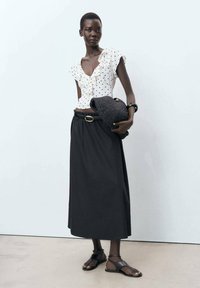 White blouse with black polka dots and ruffled detail, paired with a long black skirt. Holding a woven black handbag, wearing brown sandals.