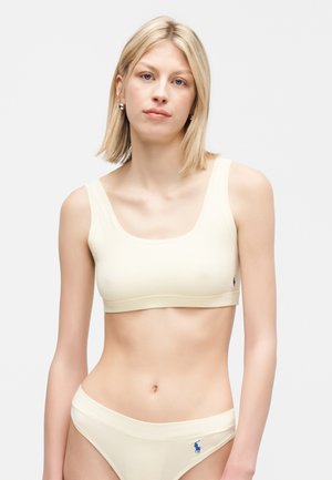 Ung kvinde med blondt hår iført cremefarvet sports-bh og matchende undertøj med lille logo på venstre side, stående mod en ensfarvet baggrund.