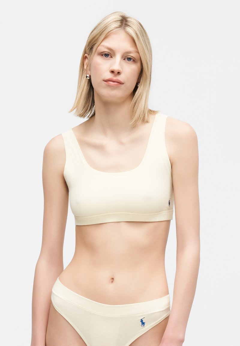 Νεαρή γυναίκα με ξανθά μαλλιά, που φοράει κορμάκι αθλητικό μπεζ και ταιριαστό εσώρουχο με μικρό λογότυπο στην αριστερή πλευρά, στέκεται μπροστά σε απλό φόντο.