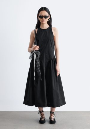 Žena v čiernych midi šatách bez rukávov, čiernych šnurovacích balerínach, čiernych slnečných okuliaroch, kruhových náušniciach a držiaca čiernu kabelku s bodkovaným šálom.