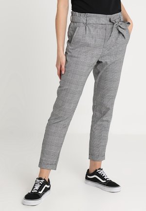 Personne portant un pantalon gris à taille paperbag à carreaux avec des poches latérales et des baskets basses noires avec des lacets et semelles blanches.