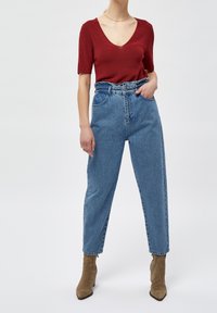 Czerwony top z krótkim rękawem i głębokim dekoltem w kształcie litery V, zestawiony z jasnoniebieskimi dżinsami o wysokim stanie oraz brązowymi zamszowymi botkami za kostkę. Teksturowany materiał na górze.